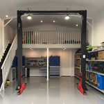 A two post car hoist with red arms and a black frame by Tufflift Hoists installed in a clean, organized workshop with blue cabinets, a workbench, shelves, and a staircase leading to a loft area. - 2 Post Clear Floor Hoist - 4.5 Ton | EE-C10V2