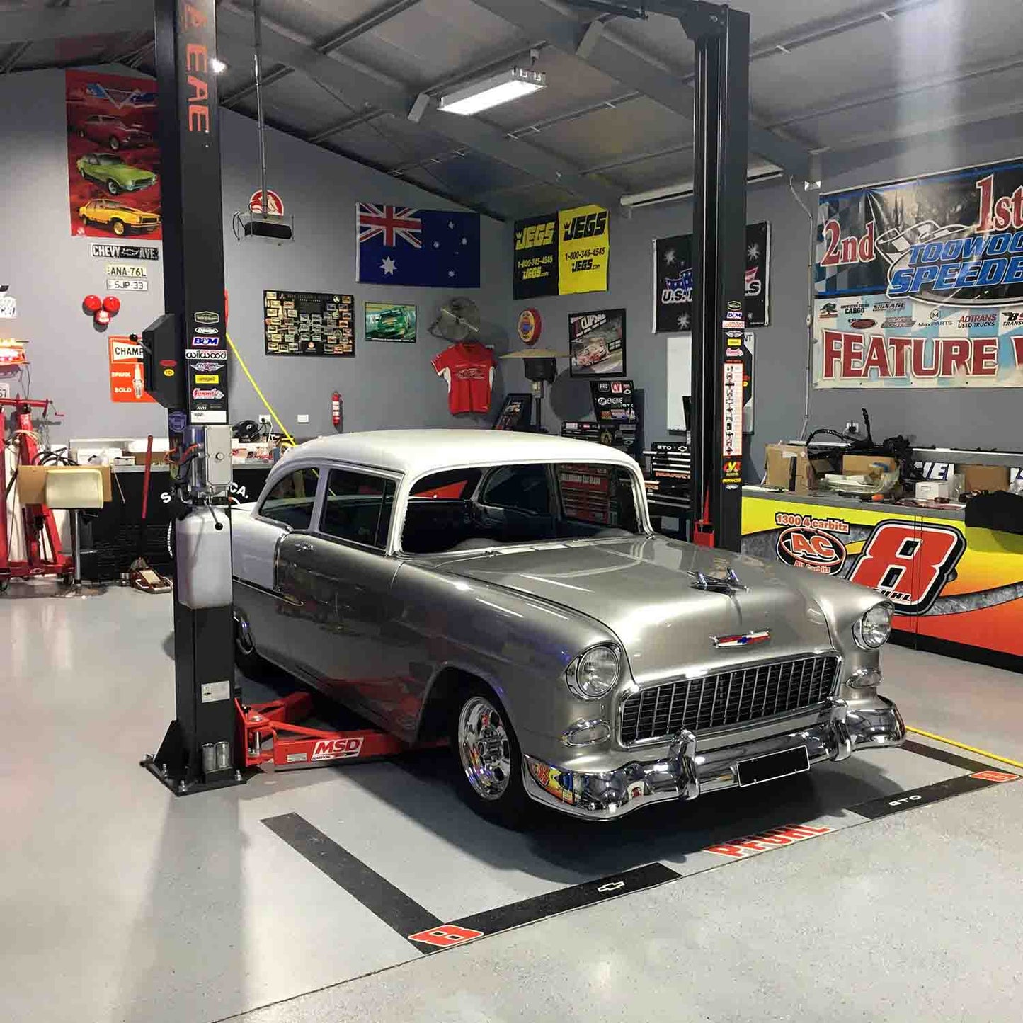 A classic car under a 2 post car hoist in a garage decked with automotive and racing decor. - 2 Post Clear Floor Hoist - 4 Ton | EE-C9