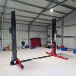 A 2 post baseplate car hoist with red arms and dark grey columns, in a well-lit garage. - 2 Post Baseplate Hoist - 4.5 Ton | EE-F10