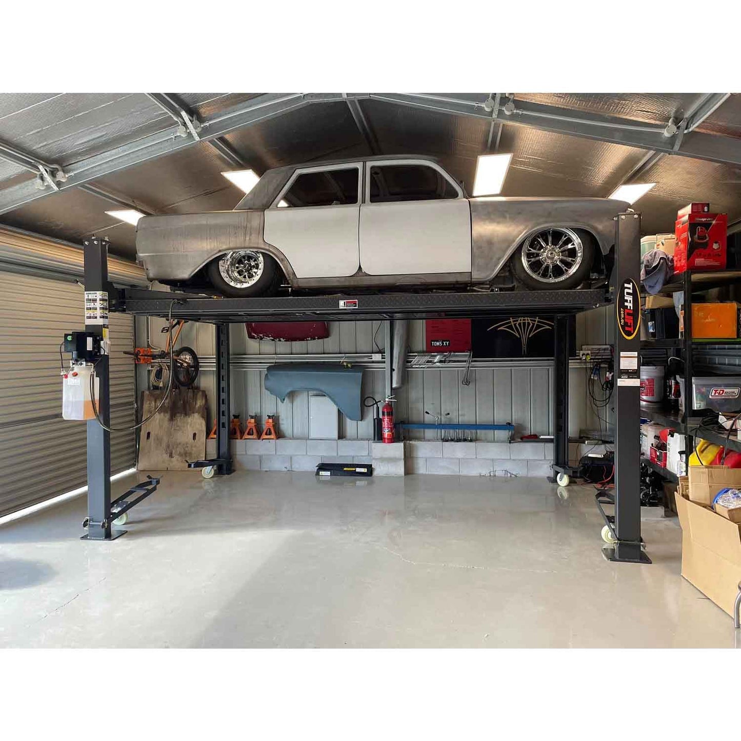 A vintage car is elevated on a 4 post parking hoist inside a well-organized garage, showcasing storage efficiency and a workspace for automotive maintenance.