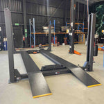 Four-post car lift with drive-on ramps installed in a modern automotive workshop, featuring tools and equipment neatly organized on shelves in the background. - 4 Post Workshop Hoist - 6 Ton | TL6.0PH