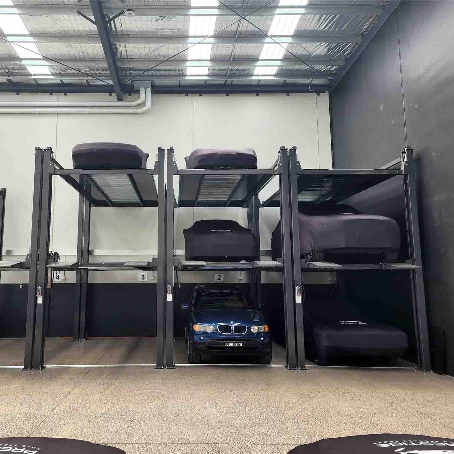 A three-tier car parking lift system in a warehouse, with a blue SUV parked on the ground level and other vehicles covered in black protective covers on the upper levels. 4 Post Parking Hoist | Triple Stacker