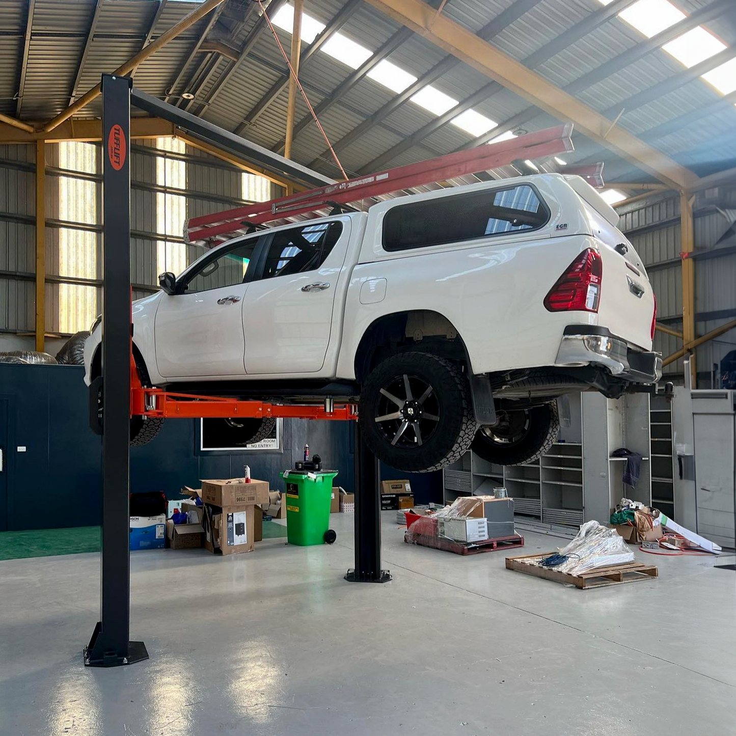 A white SUV is lifted on a hydraulic car lift inside a spacious garage or workshop. Various boxes, equipment, and tools are scattered around the floor, indicating an active workspace. The vehicle appears to be undergoing maintenance or inspection. - 2 Post Clear Floor - 5 Ton | EE-6215EKZ