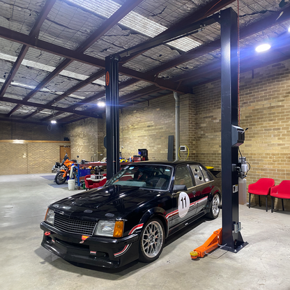 A two post car hoist with orange arms and a black frame in a brick-walled workshop, lifting a black race car with the number 11 on the door. The workshop also contains various tools, equipment, and a motorcycle in the background. - 2 Post Car Hoist - Low Profile Arms 85mm - Clear Floor - 4 Ton | EE-C9LPA