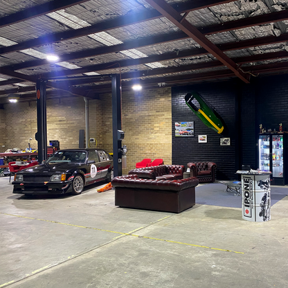 A spacious workshop with a two-post car hoist lifting a black race car numbered 11, red chairs, leather sofas, wall decorations, and a refrigerator stocked with drinks. - 2 Post Car Hoist - Low Profile Arms 85mm - Clear Floor - 4 Ton | EE-C9LPA