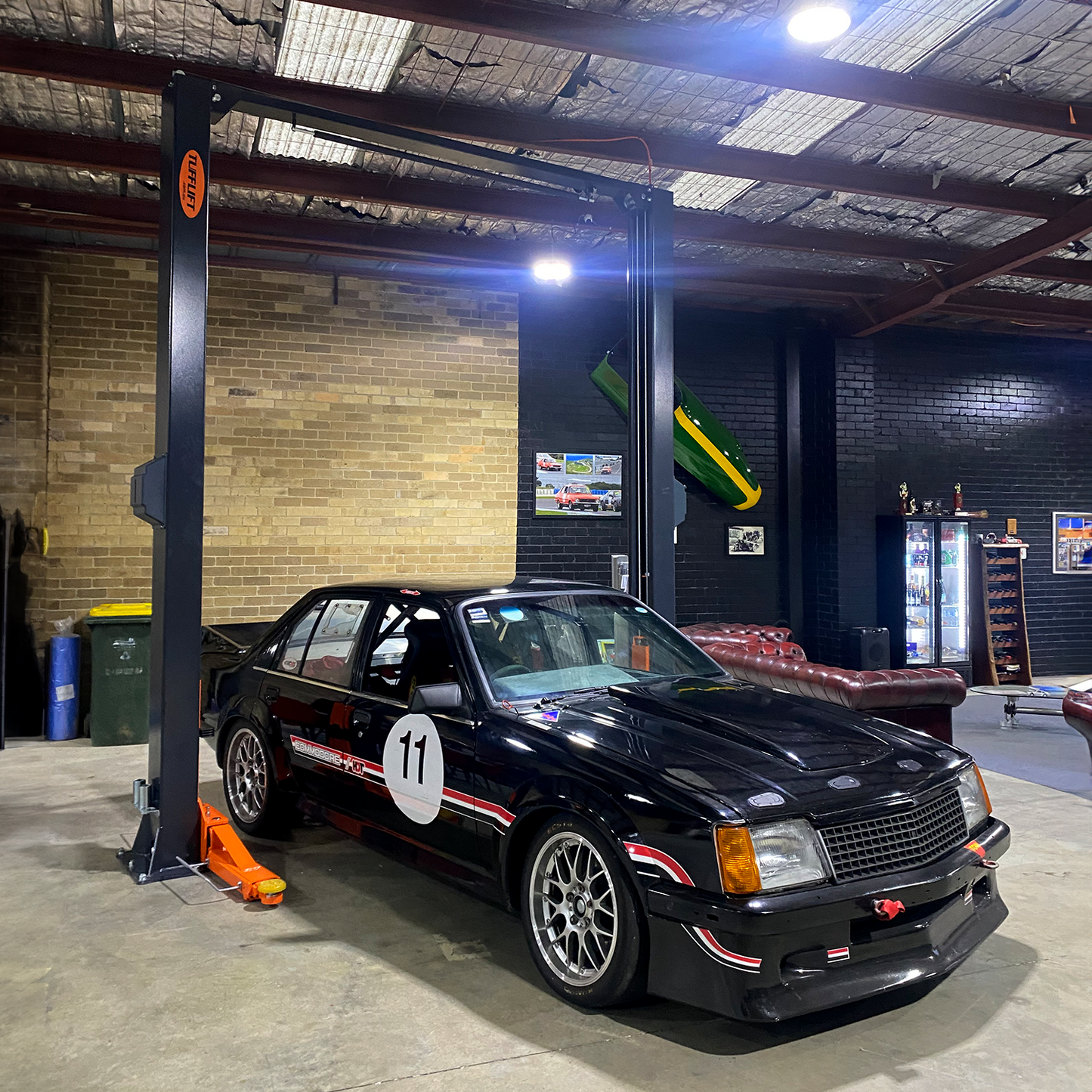 A black race car with the number 11 is positioned under a two post car hoist with orange arms in a workshop with brick walls, leather sofas, wall decorations, and a refrigerator stocked with drinks. - 2 Post Car Hoist - Low Profile Arms 85mm - Clear Floor - 4 Ton | EE-C9LPA