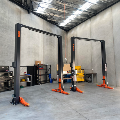 Two two-post car hoists with orange arms and black frames installed in a spacious workshop with concrete walls and various tools and equipment on shelves. - 2 Post Car Hoist - Low Profile Arms 85mm - Clear Floor - 4 Ton | EE-C9LPA