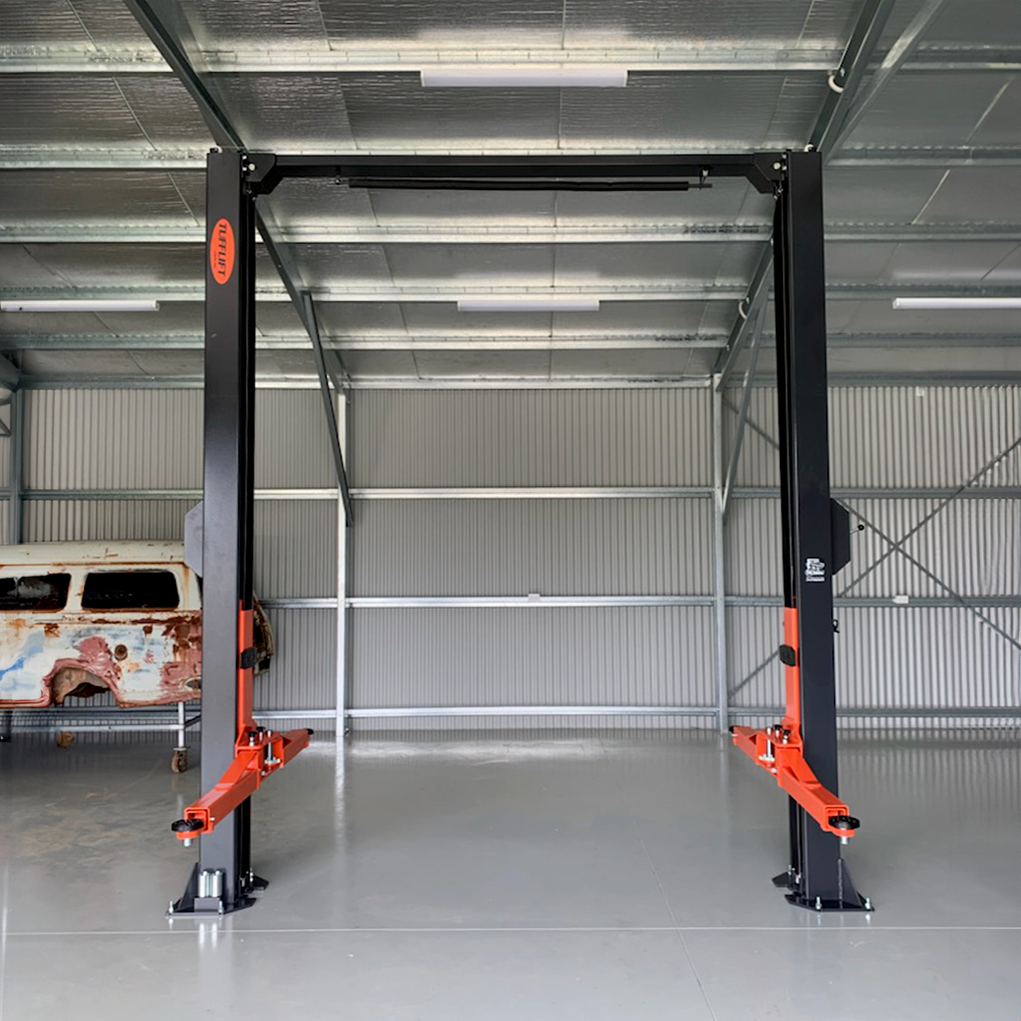 A Tufflift 2 post clear floor car hoist stands in an industrial garage, with a weathered classic car in the background. - 2 Post Clear Floor Hoist - 4 Ton | EE-C9