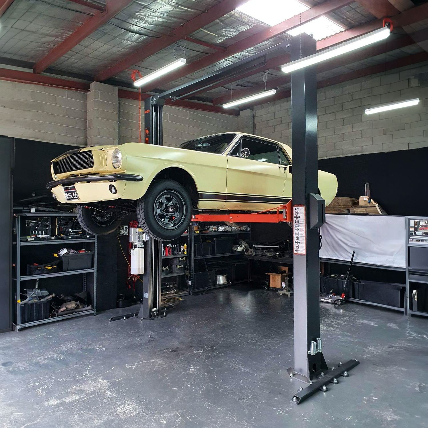 A vintage yellow Mustang is elevated on a hydraulic car hoist inside a well-organized garage. The workspace includes shelves stocked with tools and equipment, highlighting an environment dedicated to car maintenance and restoration. - 2 Post Clear Floor Hoist - 4 Ton | EE-C9