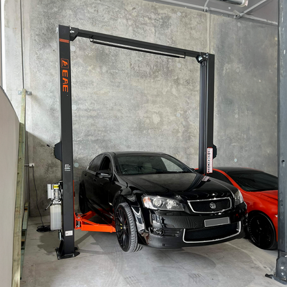 A black sedan is parked under a hydraulic car hoist inside a bare, concrete-walled garage. The car lift is not in use, and the vehicle appears to be positioned for potential maintenance or storage. A red car is partially visible next to the black sedan. - 2 Post Clear Floor Hoist - 4 Ton | EE-C9