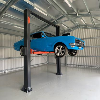 A vibrant blue classic car is elevated on a 2 post clear floor car hoist in a well-lit metal shed, showcasing the vehicle's undercarriage. - 2 Post Clear Floor Hoist - 4 Ton | EE-C9