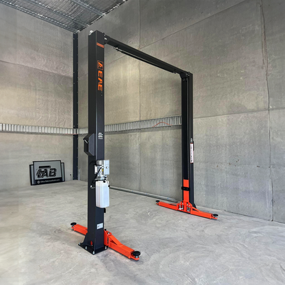 A hydraulic car lift stands idle in an empty garage with bare concrete walls. The lift's arms are spread out, ready to accommodate a vehicle. A framed sign is propped against the wall in the background. - 2 Post Clear Floor Hoist - 4 Ton | EE-C9