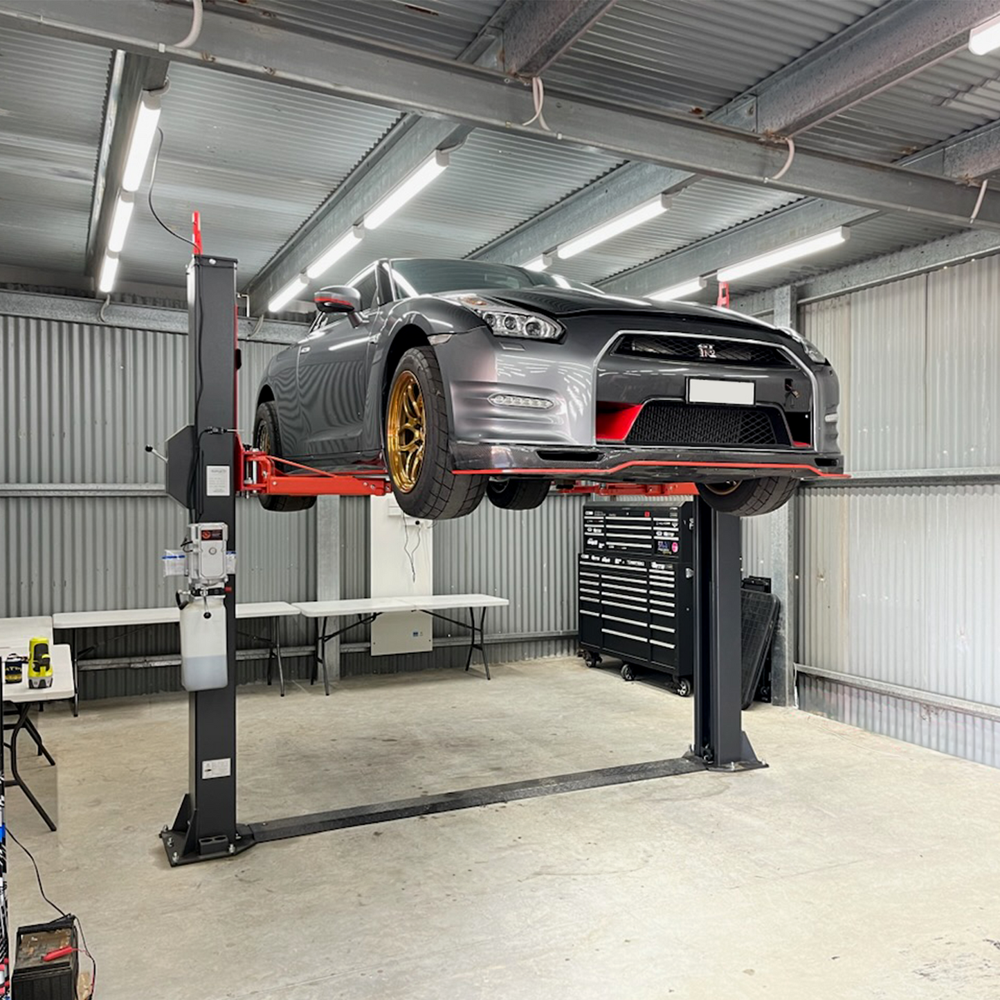 A sleek car with golden wheels is elevated on a red and black Tufflift 2 post baseplate car hoist in an industrial garage, with workbenches and tools in the background, ready for maintenance or repair. - 2 Post Baseplate Hoist - 4.5 Ton | EE-F10