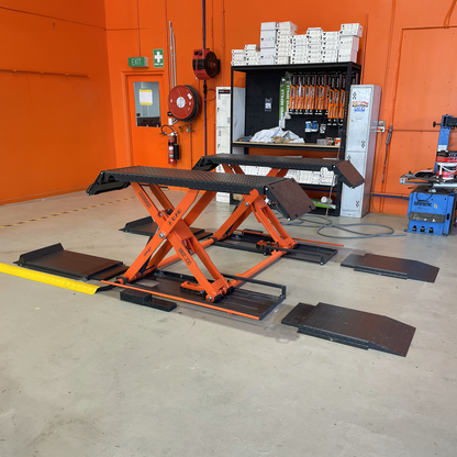 An orange EAE hydraulic scissor lift, model MR35, in a workshop setting. The lift is in a raised position with its ramps and platforms visible. The surrounding workspace includes shelves with tools and equipment, an orange wall, and various safety equipment.