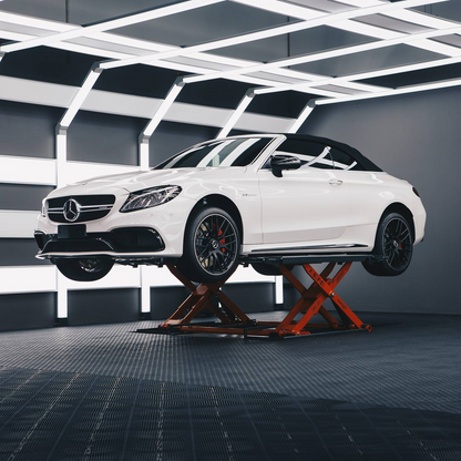 A white Mercedes-Benz AMG convertible is elevated on an orange EAE hydraulic scissor lift, in a modern, well-lit garage. The sleek lighting and clean environment emphasize the high-end automotive service setting.