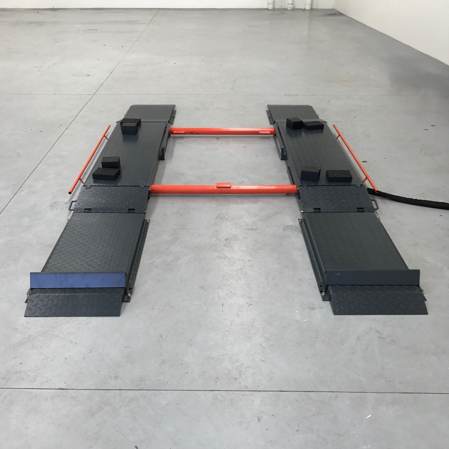 Scissor lift setup with approach ramps and orange crossbars, positioned on a clean concrete floor. The setup includes rubber chocks on the platforms, providing additional support and stability for SUV or 4WD lifting.