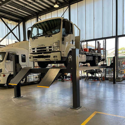 Isuzu FRR 500 truck elevated on a 12,000 kg capacity 4 post truck hoist inside a modern industrial garage, with bright lighting and other commercial vehicles visible in the background. The hoist features yellow-edged diamond-plated ramps and a pneumatic single-point lock release. - 4 Post 7.5M Long Truck Hoist – 12 Ton | TL12.0TH-7.5