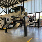 Isuzu FRR 500 truck elevated on a 12,000 kg capacity 4 post truck hoist inside a modern industrial garage, with bright lighting and other commercial vehicles visible in the background. The hoist features yellow-edged diamond-plated ramps and a pneumatic single-point lock release. - 4 Post 7.5M Long Truck Hoist – 12 Ton | TL12.0TH-7.5