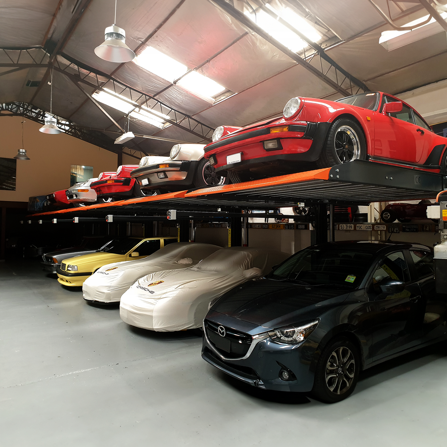 Indoor parking garage featuring a multi-level 2 post car hoist system, with classic red sports cars on the upper platform and a row of covered vehicles including a yellow car, two white-covered cars, and a dark blue car on the ground level. - 2 Post Parking Hoist - Full Platform Compact - 2.7 Ton | TL2.7HP