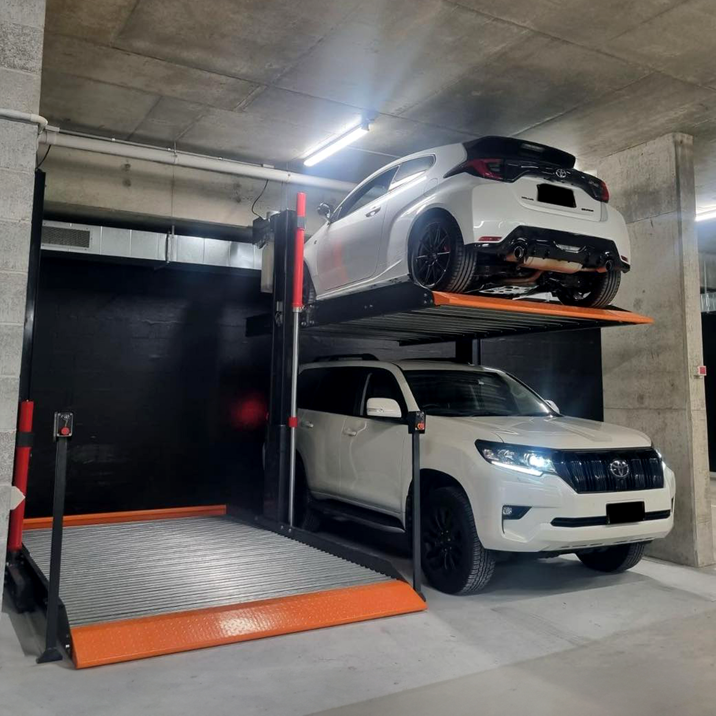 Platform 2 post car hoist system in a concrete-walled garage, with a white SUV lifted above another white SUV, demonstrating the hoist's capability for efficient vertical vehicle storage. - 2 Post Parking Hoist - Full Platform Compact - 2.7 Ton | TL2.7HP