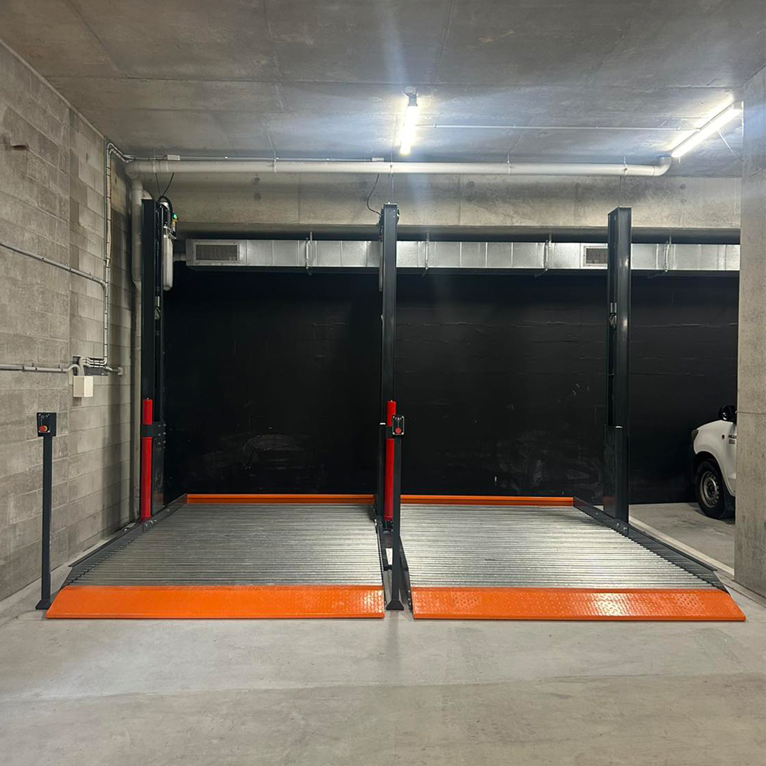 Empty platform 2 post car hoist system in a concrete-walled garage, featuring wide platforms with orange safety accents, ready for vehicle lifting. - 2 Post Parking Hoist - Full Platform Compact - 2.7 Ton | TL2.7HP