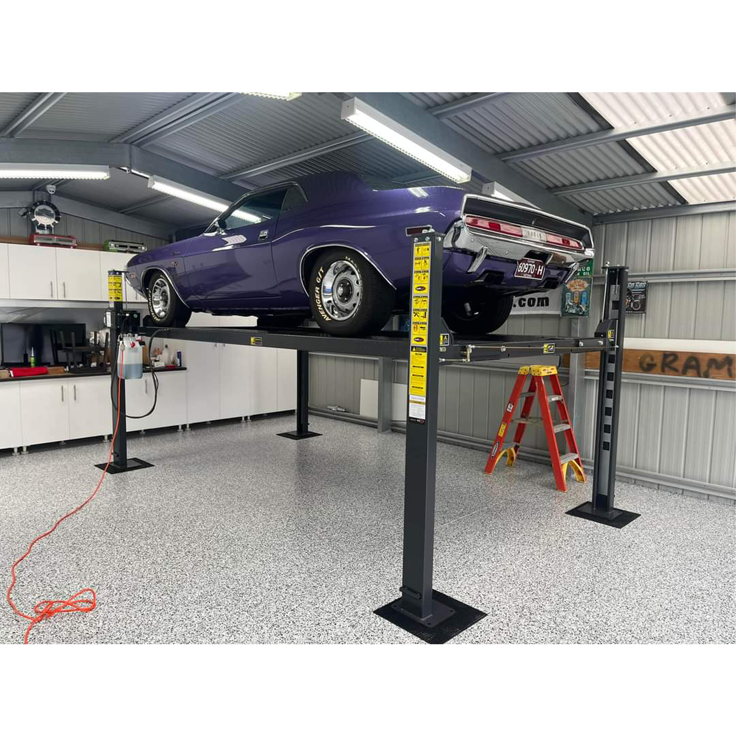 A gleaming purple vintage muscle car raised on a 4 post parking hoist, with a hydraulic power unit visible on the left; the garage is clean and modern with white cabinets and speckled flooring.