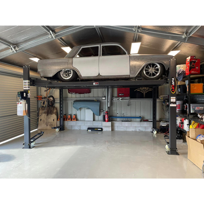 A vintage car is elevated on a 4 post parking hoist inside a well-organized garage, showcasing storage efficiency and a workspace for automotive maintenance.