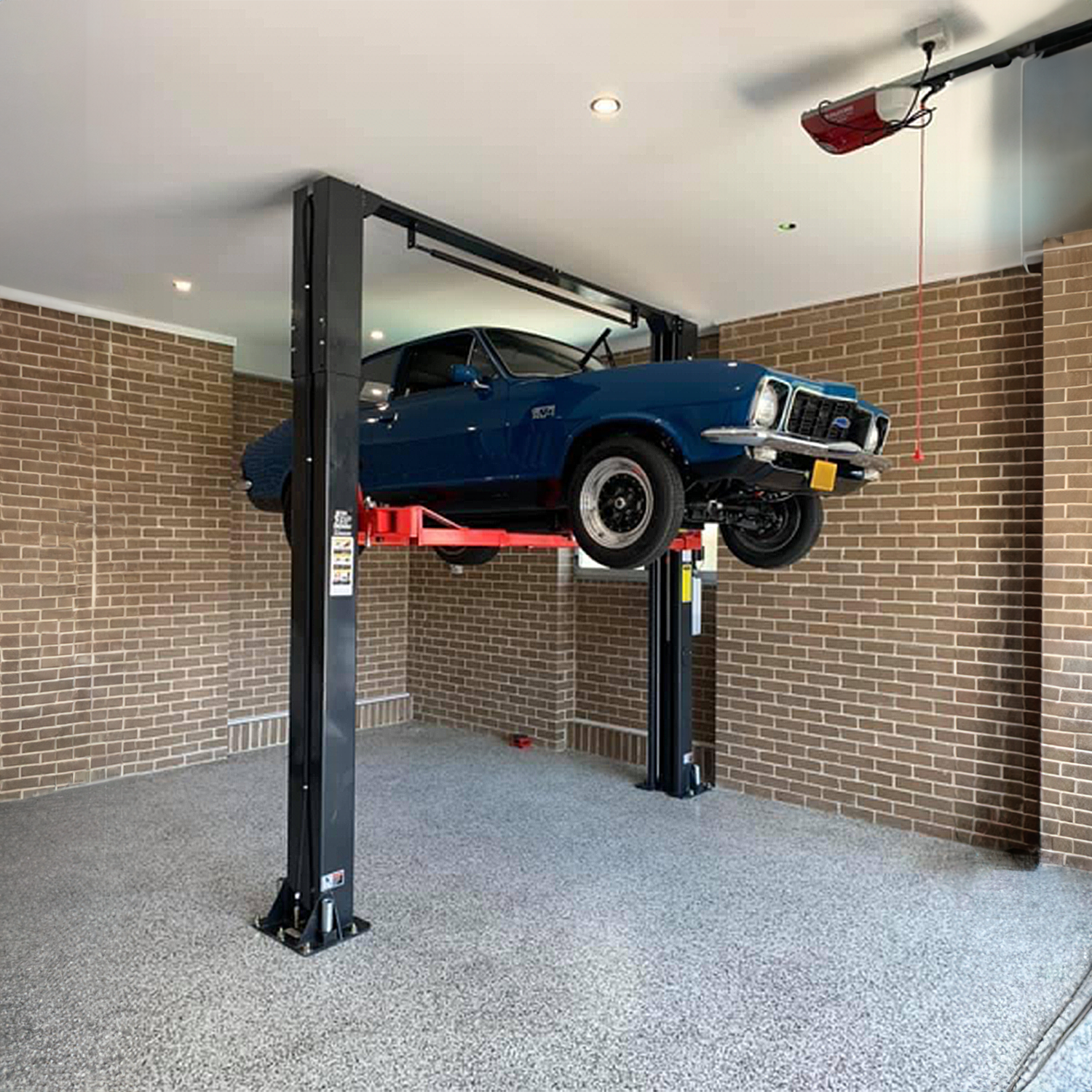 A classic blue muscle car is elevated on a dark dry and red Tufflift clear floor car hoist in a well-lit garage with brick walls. The car is lifted high above the ground. - 2 Post Clear Floor Hoist - 4 Ton | TL4.0OHDI