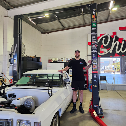 Charlie Dixon, wearing a black shirt and shorts, stands proudly next to a white vintage car with its hood removed, revealing a turbocharger inside. The car is positioned on a 2 post car hoist in a well-lit garage with a Tufflift and various car brand stickers on the lift column. - 2 Post Clear Floor Hoist - 4 Ton | TL4.0OHDI