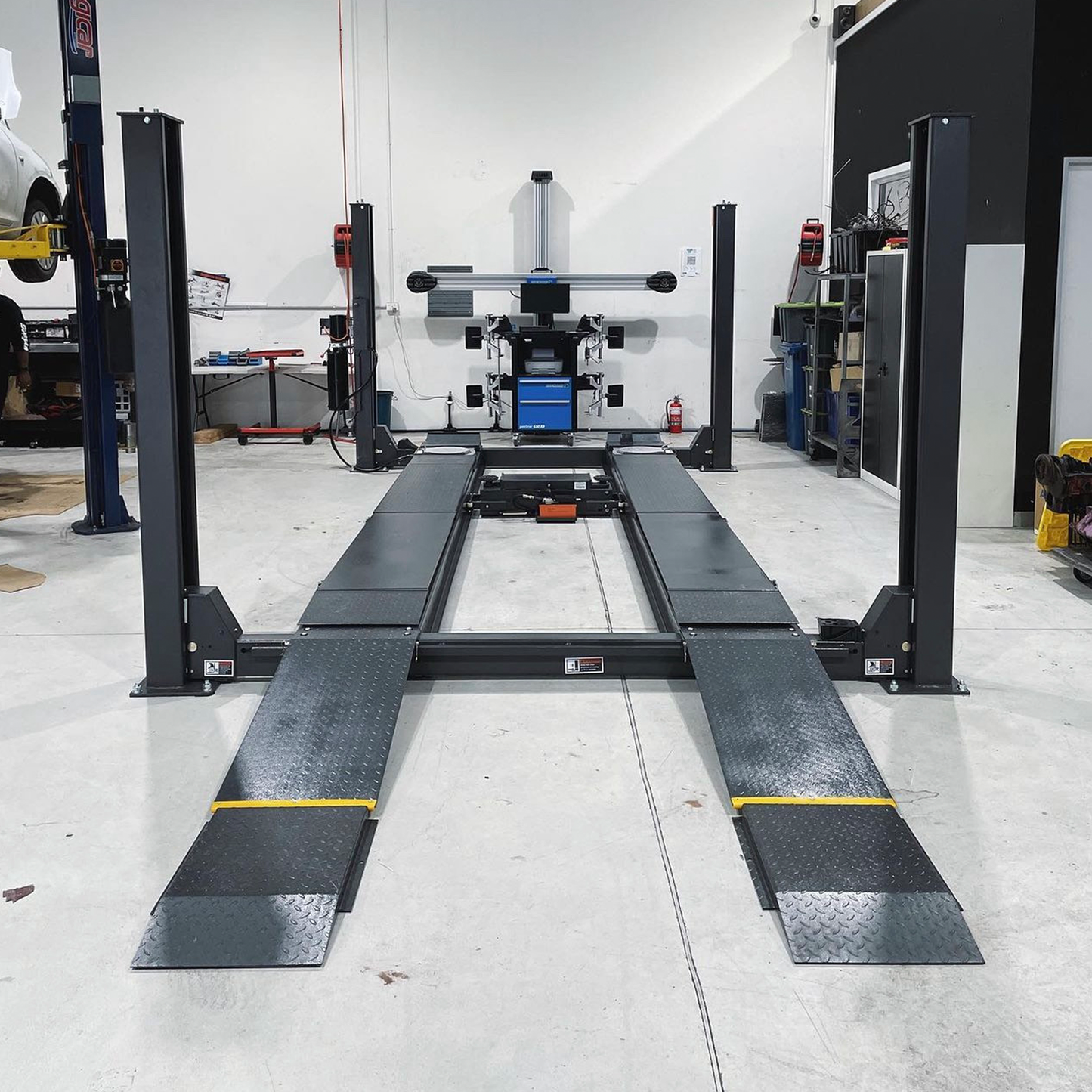 A four-post wheel alignment hoist in a workshop, featuring ramps and a wheel aligner machine in the background for precise vehicle alignment and maintenance. - 4 Post Wheel Alignment Hoist - 6 Ton | TL6.0PH-WA